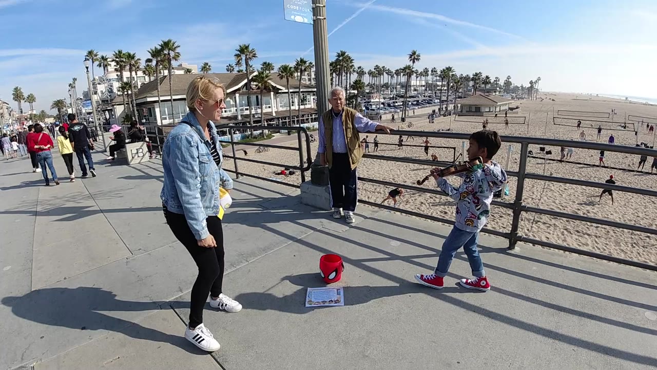 At Huntington Beach, a child plays violin near the railing while a woman stands close in the foreground and an older man watches from behind.