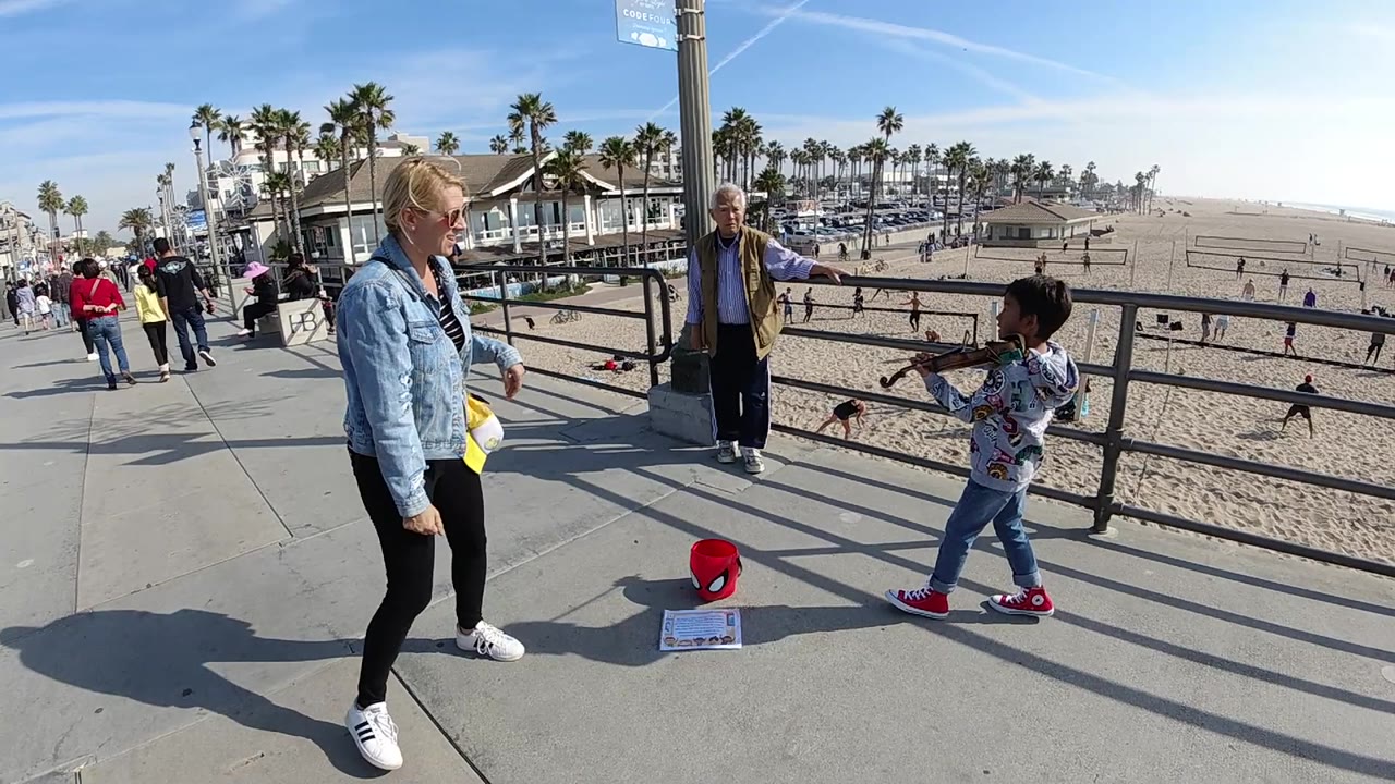 At Huntington Beach, the child holds the violin and bow near the railing as the woman shifts position and beachgoers move in the distant sand and walkway.