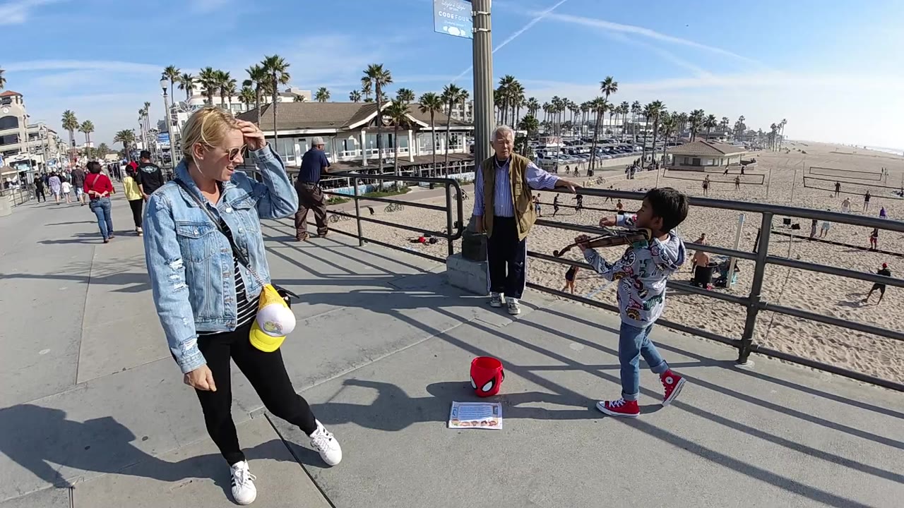 At 0:00 in Huntington Beach, California, a child plays violin near a railing while a woman in sunglasses walks in the foreground and beachgoers move across the sand behind them.