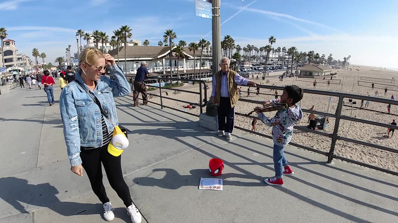 At 0:02 in Huntington Beach, California, the child violinist holds the bow near the right side of the frame as the foreground woman looks downward and the sandy beach stretches into bright haze.