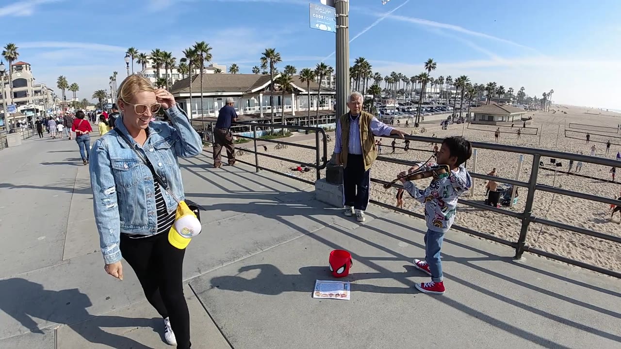 Beach promenade scene with child violinist, passersby, railing, sand, and palm trees.