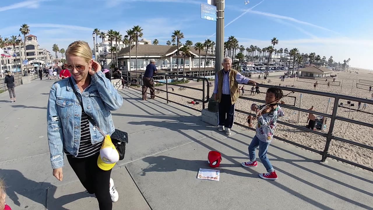 At 0:02 in Huntington Beach, California, the woman is slightly farther through the foreground while the child, older man, railing, sandy beach, palms, and marina remain visible behind her.