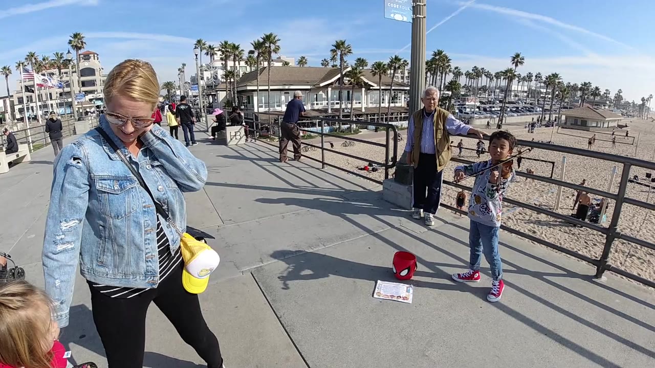 At Huntington Beach, a child holds a violin beside a railing as a woman in a denim jacket looks down nearby, with sand, palms, pedestrians, and waterfront buildings behind them.