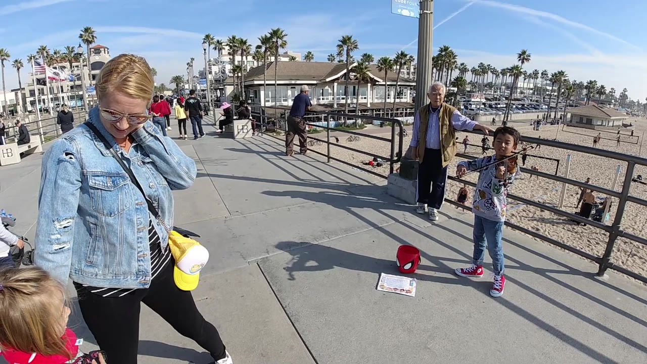 At Huntington Beach, the child violinist stands on the right side of the walkway as a woman and a small child occupy the foreground, with an older man, railings, sand, palms, and marina-side buildings visible beyond.