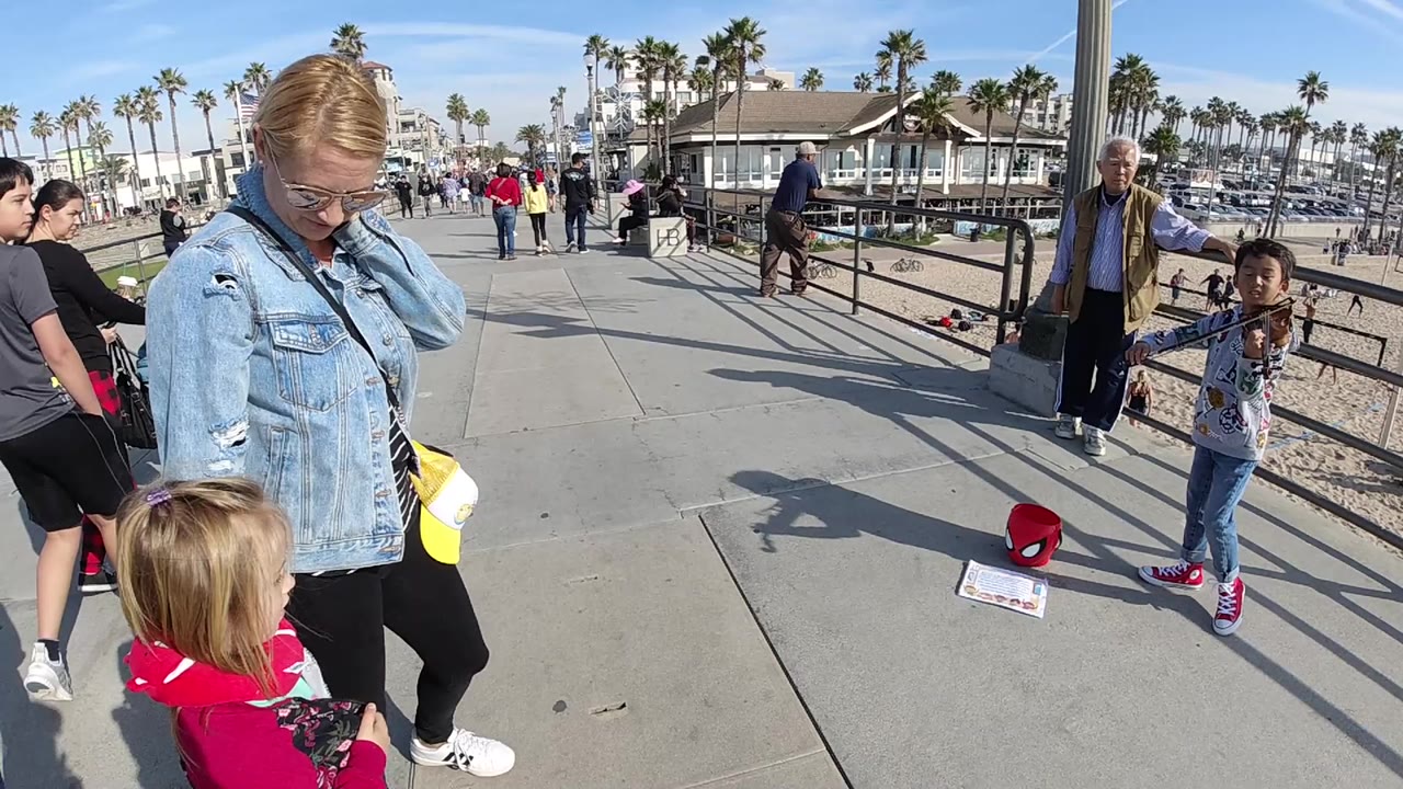 In Huntington Beach, a woman in a denim jacket and a small child stand near a railing as a young violinist performs on a sunny sidewalk with pedestrians behind them.