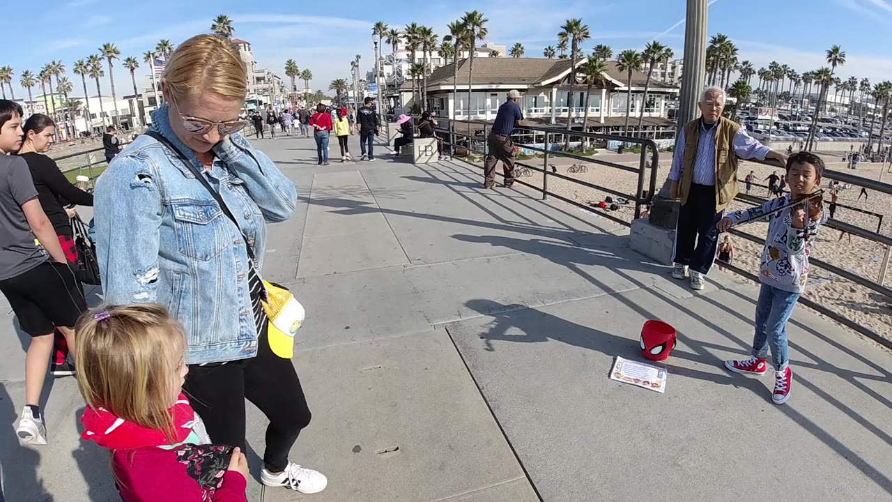 In Huntington Beach, the small child in red remains in the foreground while the woman looks down and the young violinist stands near a red mask on the pavement.
