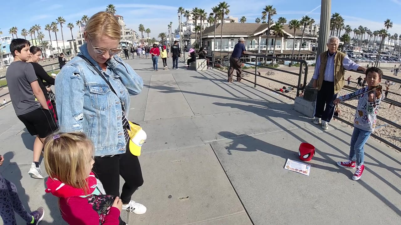 In Huntington Beach, the woman in denim and the child in red stand in the foreground while a young violinist and several pedestrians occupy the palm-lined sidewalk behind them.