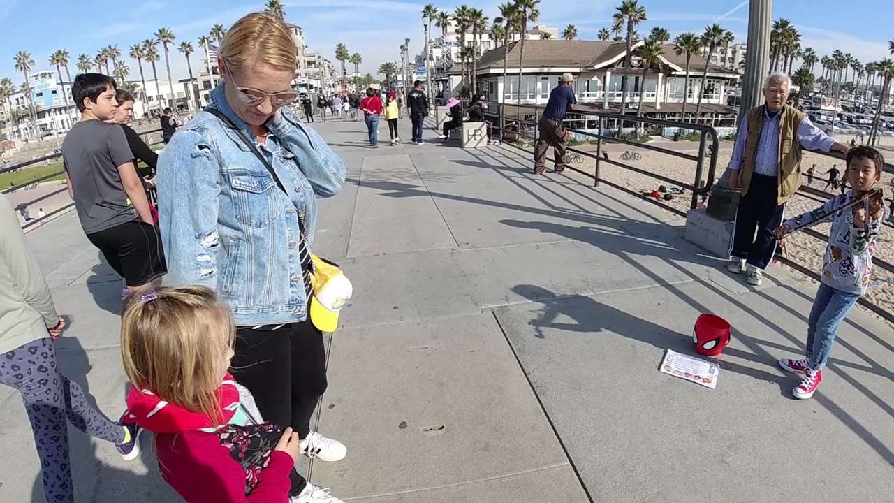 A girl walks between the camera and a woman watching a young violinist.