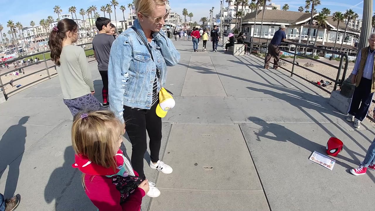 A woman in a denim jacket stands near a young child in pink on a Huntington Beach walkway, with railings, palm trees, beachgoers, and long shadows in view.