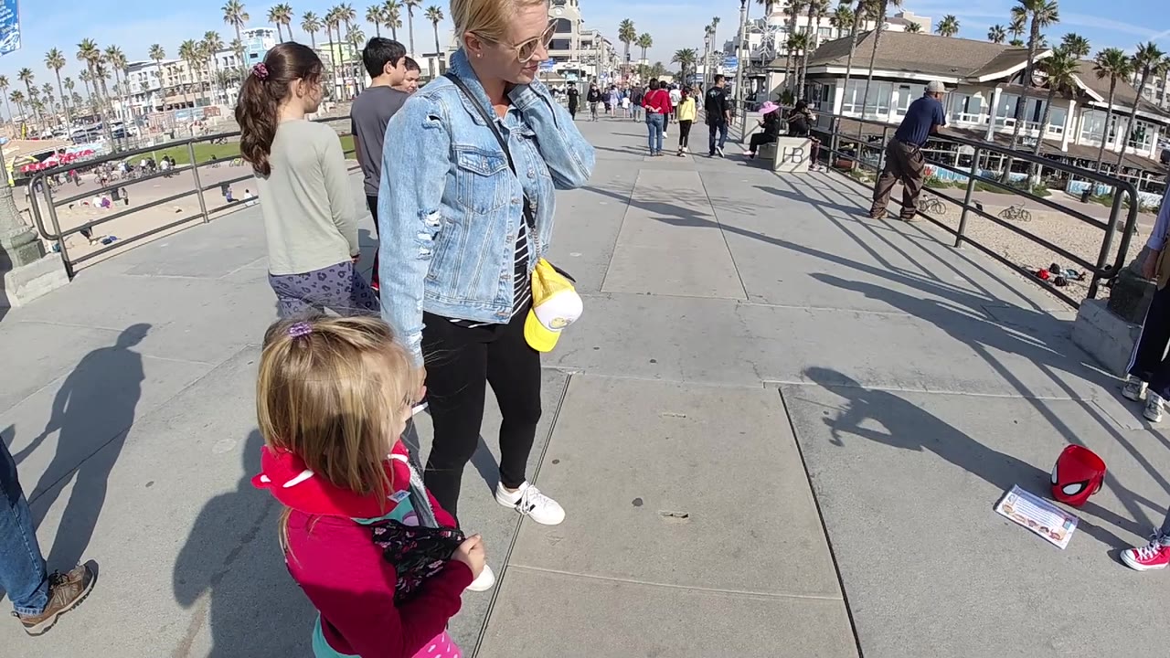 A wide-angle view shows the woman and child near the lower edge of the Huntington Beach walkway while pedestrians pass through the sunlit midground.
