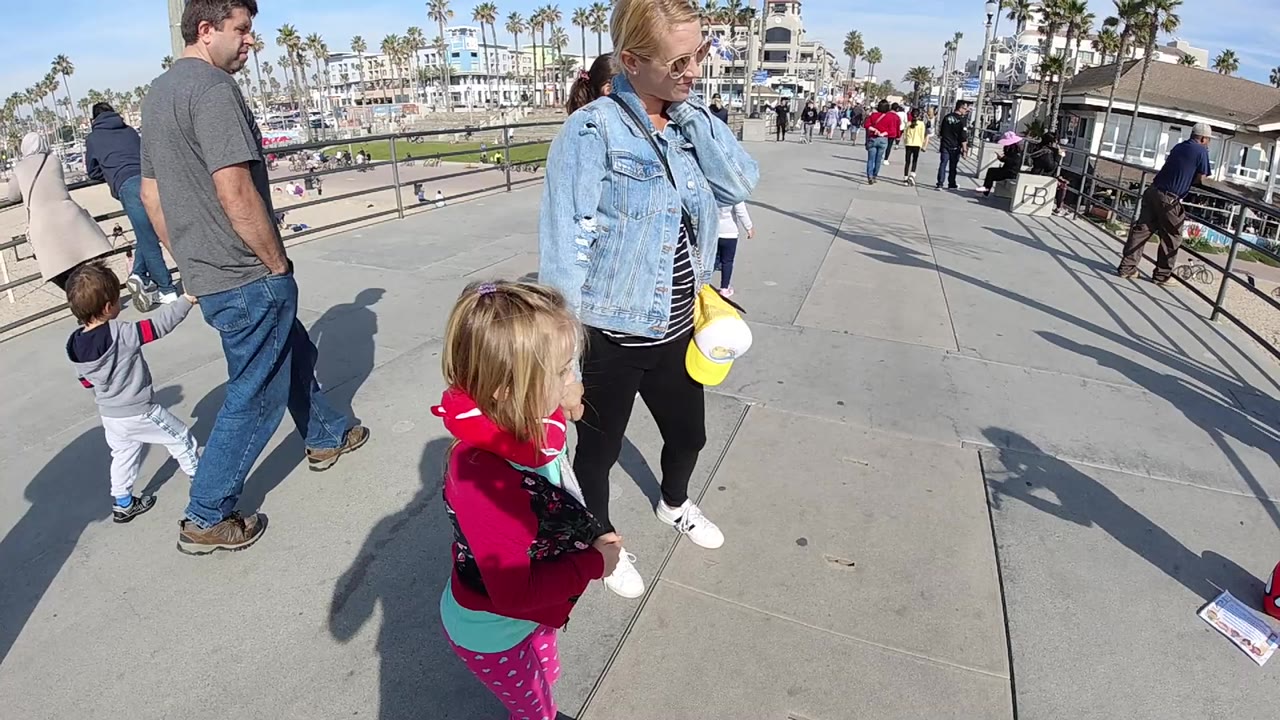 A wide close view of adults and children standing on a paved Huntington Beach walkway, with railings, palm trees, beachfront buildings, blue sky, and long shadows visible.