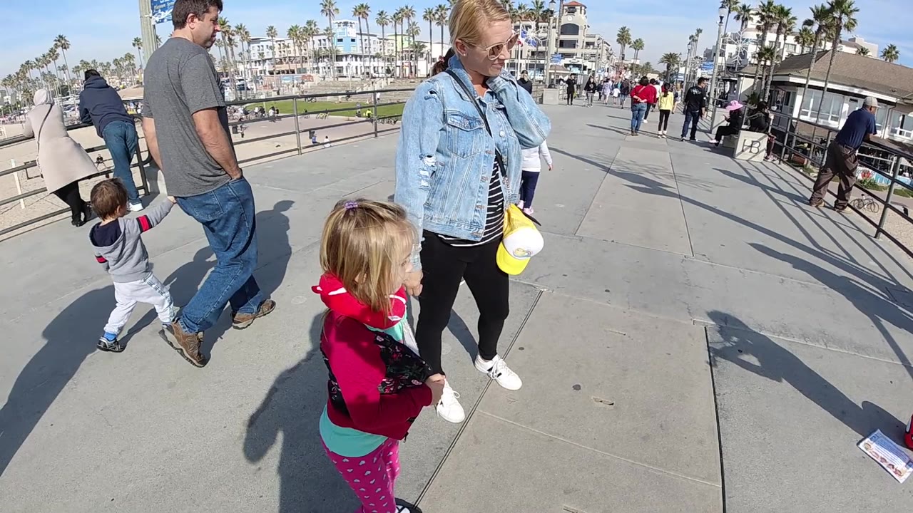 The foreground children shift position beside the adults on the Huntington Beach walkway, with a railing, passing pedestrians, palm trees, and white buildings in the bright background.