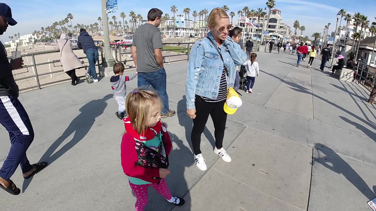 A woman in a denim jacket stands near a small child in pink clothing on a Huntington Beach boardwalk, with pedestrians, railings, sand, palm trees, and buildings behind them.
