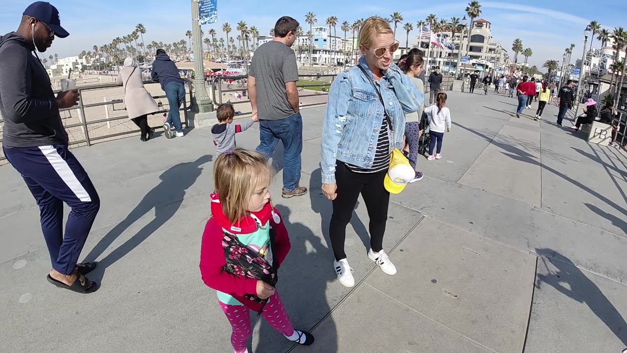 A man in dark athletic clothing walks through the left foreground as the woman and child remain near the center of the Huntington Beach boardwalk, with children and adults ahead.