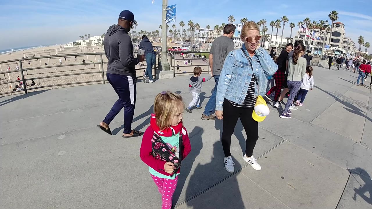 In Huntington Beach, California, a child in a red jacket stands in the foreground while a blonde woman in sunglasses and denim walks nearby beside a beach railing.