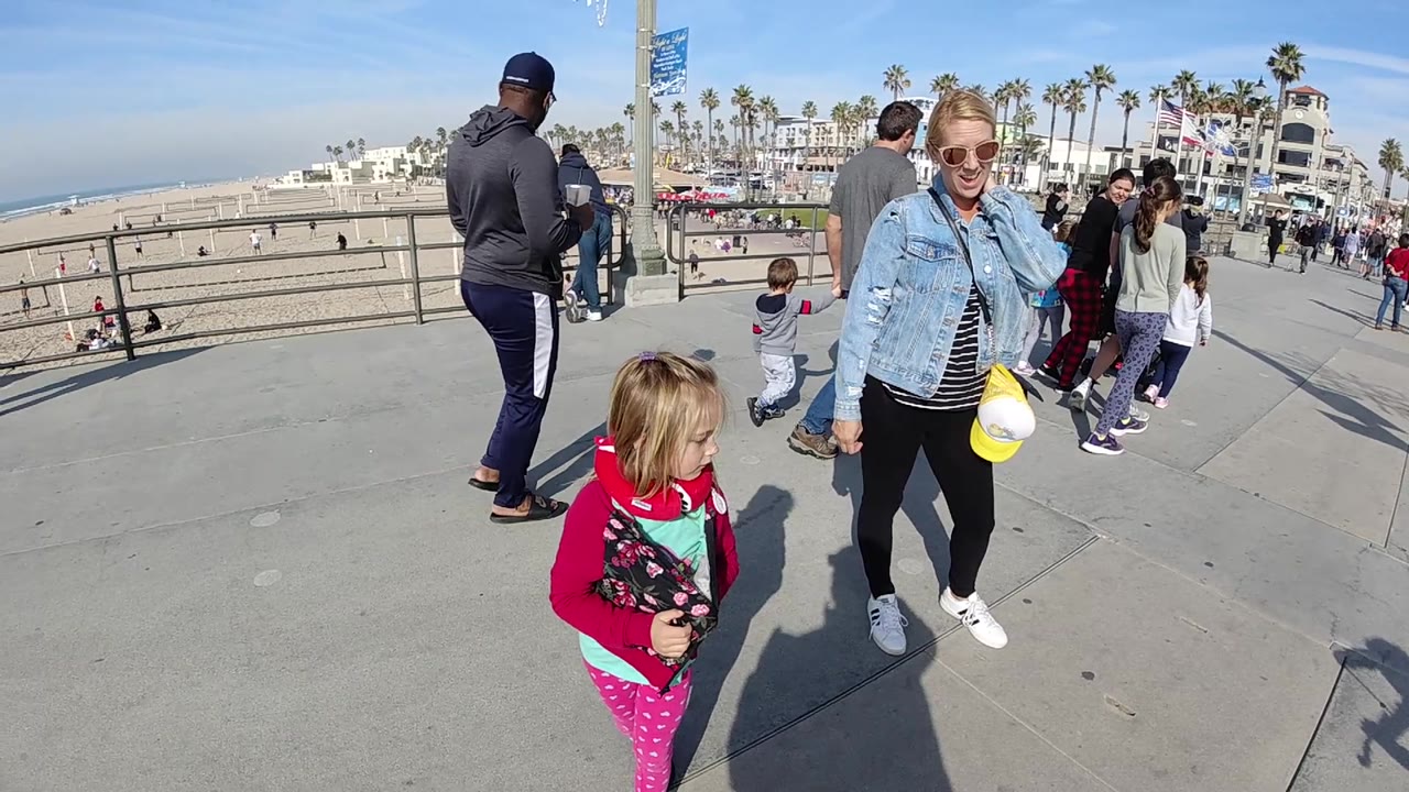 In Huntington Beach, California, the blonde woman appears closer to center while the child in red looks across the walkway, with beach, railing, pedestrians, and palms behind them.