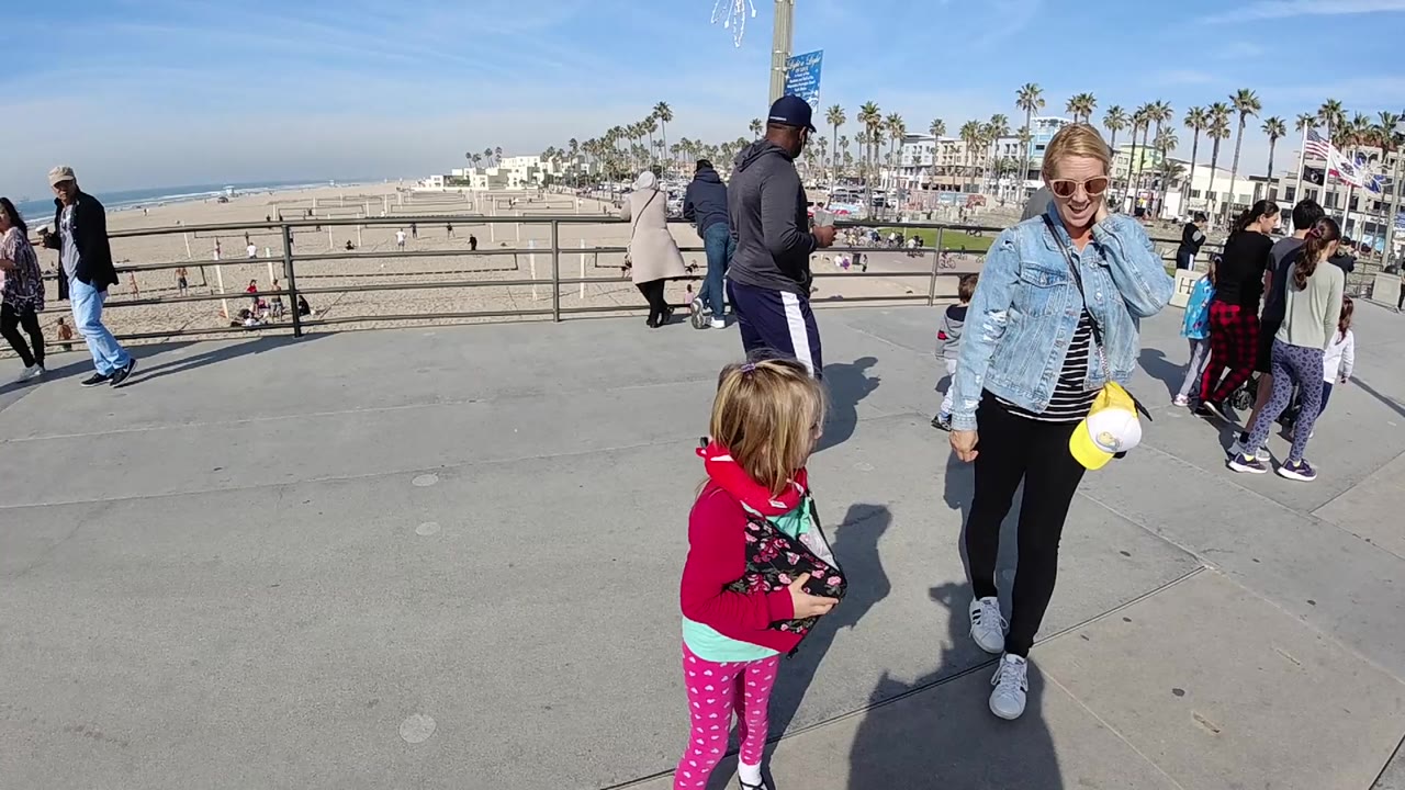 At 0:00 in Huntington Beach, a woman in a denim jacket and sunglasses stands near a young girl in a red jacket on a concrete walkway above the sand, with pedestrians and palm trees behind them.