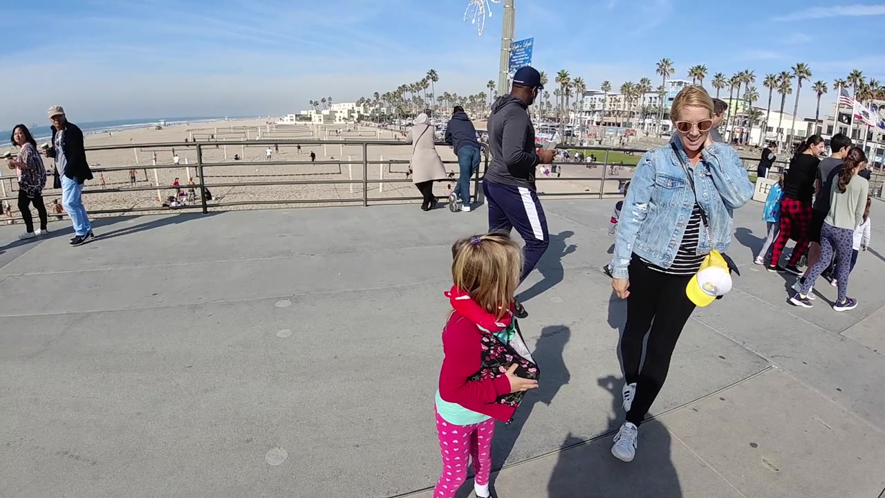 At 0:02 in Huntington Beach, the child in a red jacket stands in the foreground while the woman remains to the right, with a man in athletic clothing and other pedestrians crossing behind them.