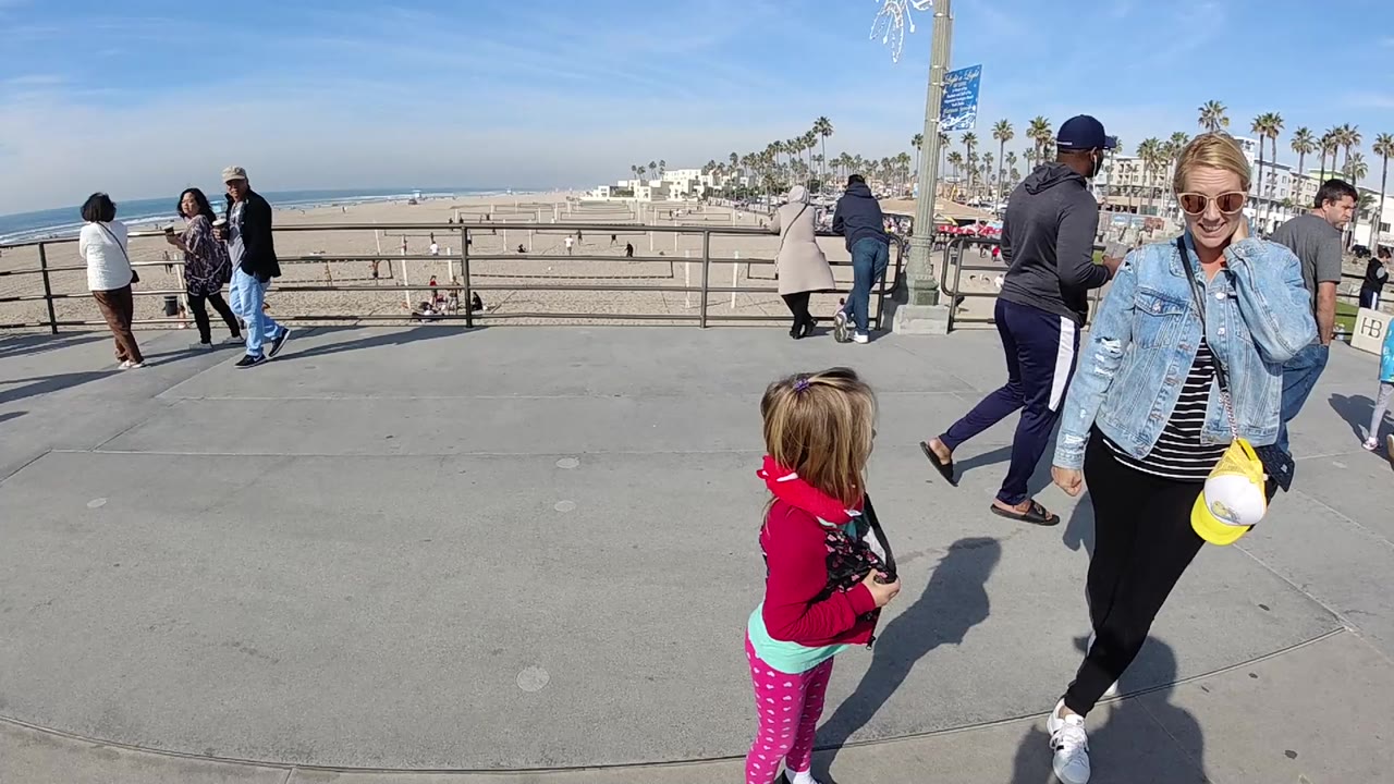 At the start of the clip, a woman in sunglasses and a denim jacket walks near a young child on a Huntington Beach promenade with sand, ocean, railings, palm trees, and pedestrians behind them.