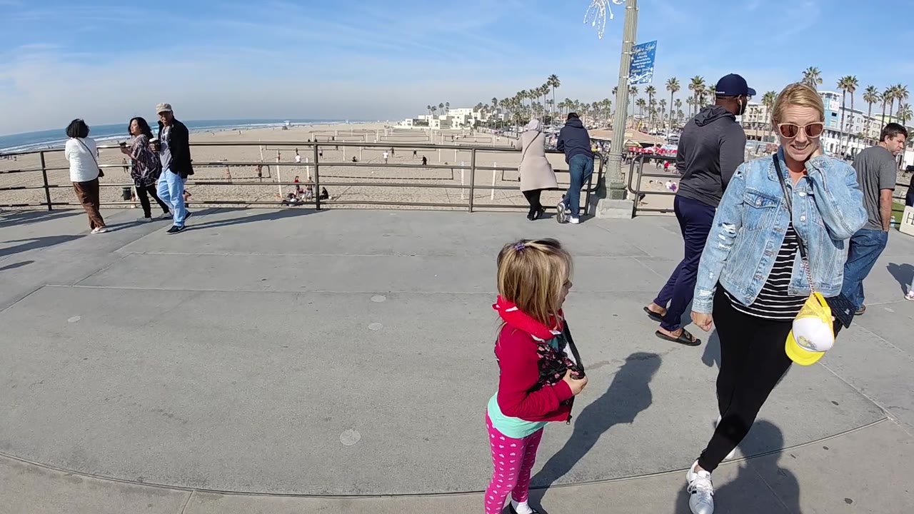 At 0:02, the woman and child remain in the foreground on the Huntington Beach promenade, with strong shadows on the concrete and palm-lined buildings along the beach in the distance.