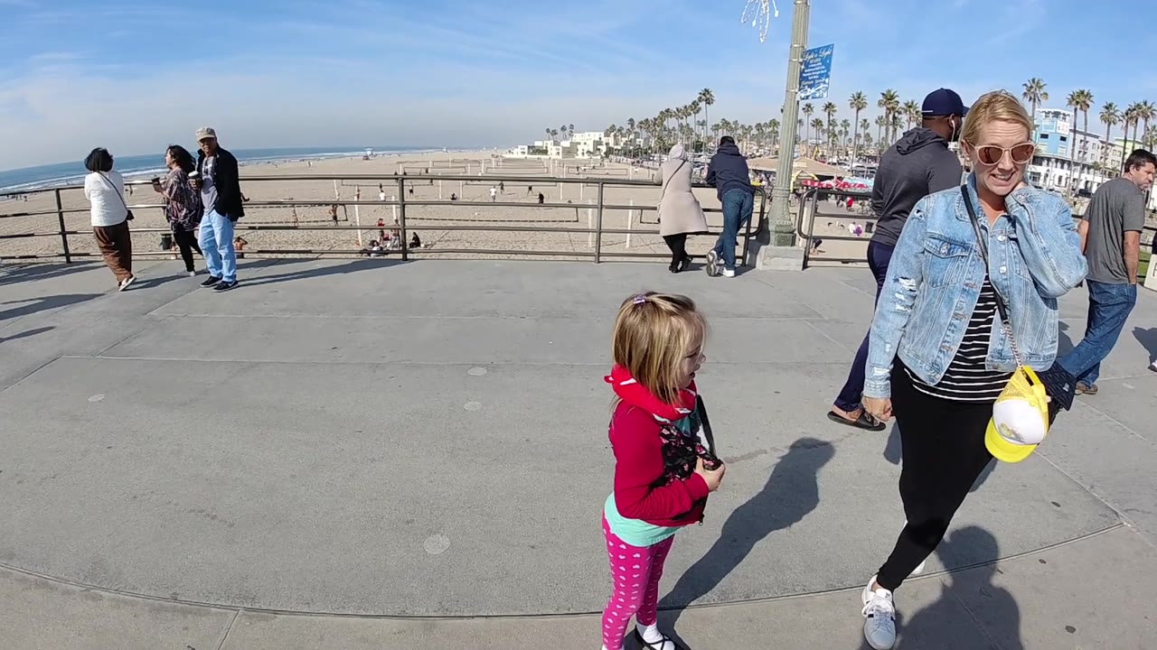 Child in pink stands near a woman on a beach promenade.