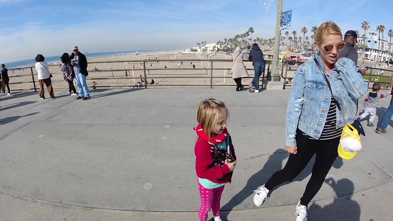 At 0:02, the child in a red hoodie stands closer to frame center while the woman with sunglasses and a yellow item remains beside them on the Huntington Beach boardwalk.