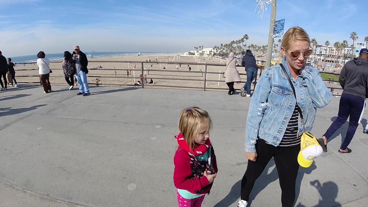 At Huntington Beach, a woman in sunglasses and a denim jacket stands near a child in a pink hoodie on a concrete promenade above the sand and ocean.