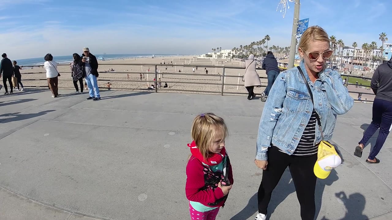 At Huntington Beach, the woman is partly cropped at the right edge while the child stands lower in frame, with broad concrete paving, railings, beach, ocean, and palm trees visible.