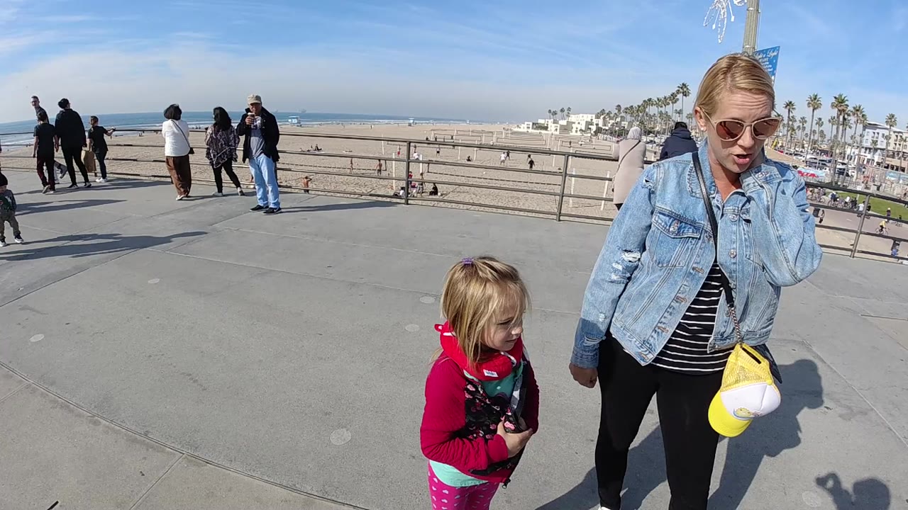 At 0:00 in Huntington Beach, California, a woman in sunglasses and a denim jacket walks beside a young girl in pink on a concrete boardwalk above the beach.