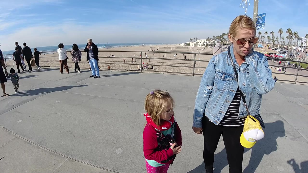 At 0:00 in Huntington Beach, a woman in sunglasses and a denim jacket stands beside a young child in red near a railing, with beach pedestrians and ocean visible behind them.
