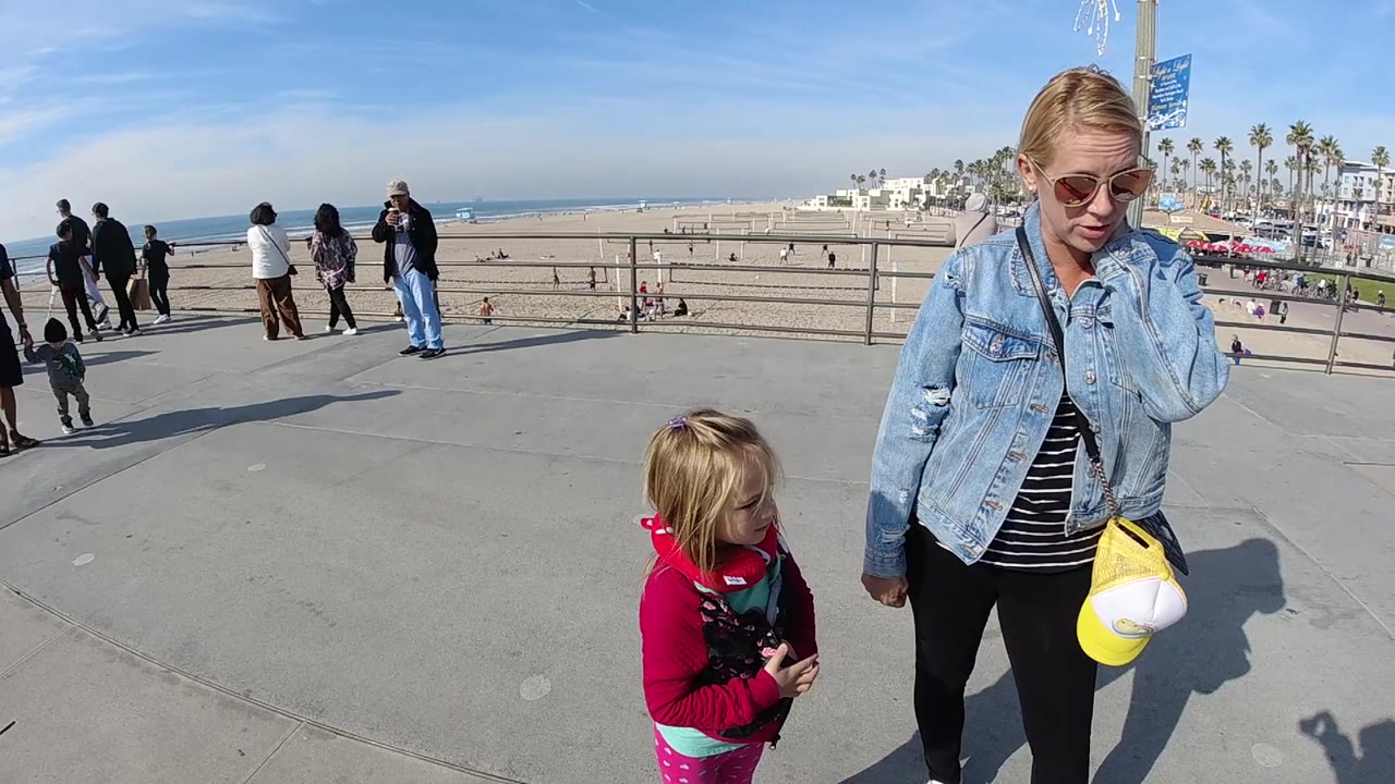At 0:02 in Huntington Beach, the woman looks downward beside the child in red, with the beach railing, sandy shore, ocean line, and palm trees across the background.