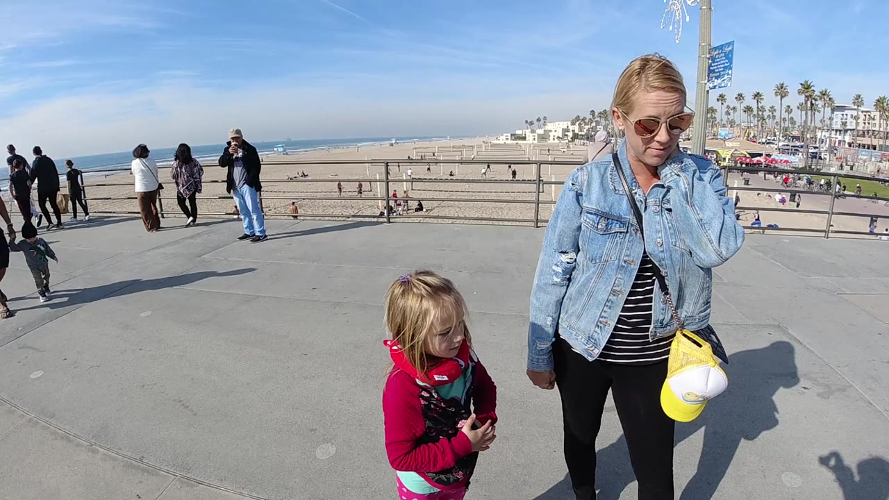 At 0:00, a woman in sunglasses and a denim jacket stands beside a young child on a paved overlook above the Huntington Beach sand and ocean.