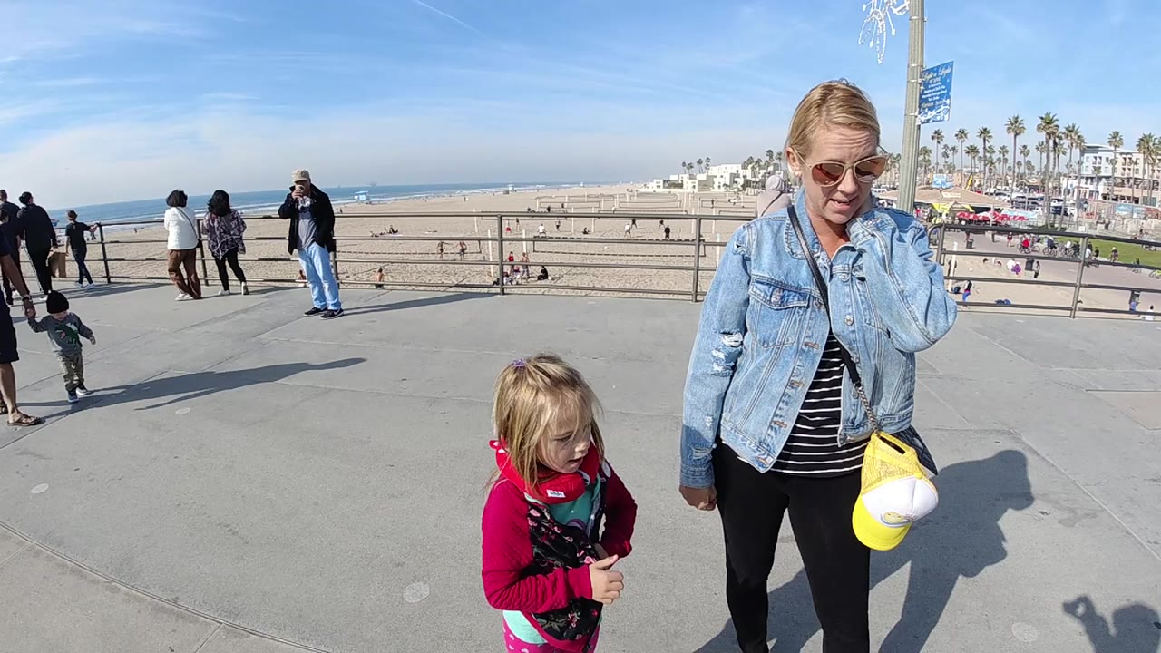 At 0:00 in Huntington Beach, California, a woman in sunglasses and denim stands beside a young child in pink on a paved overlook above the sand and ocean.
