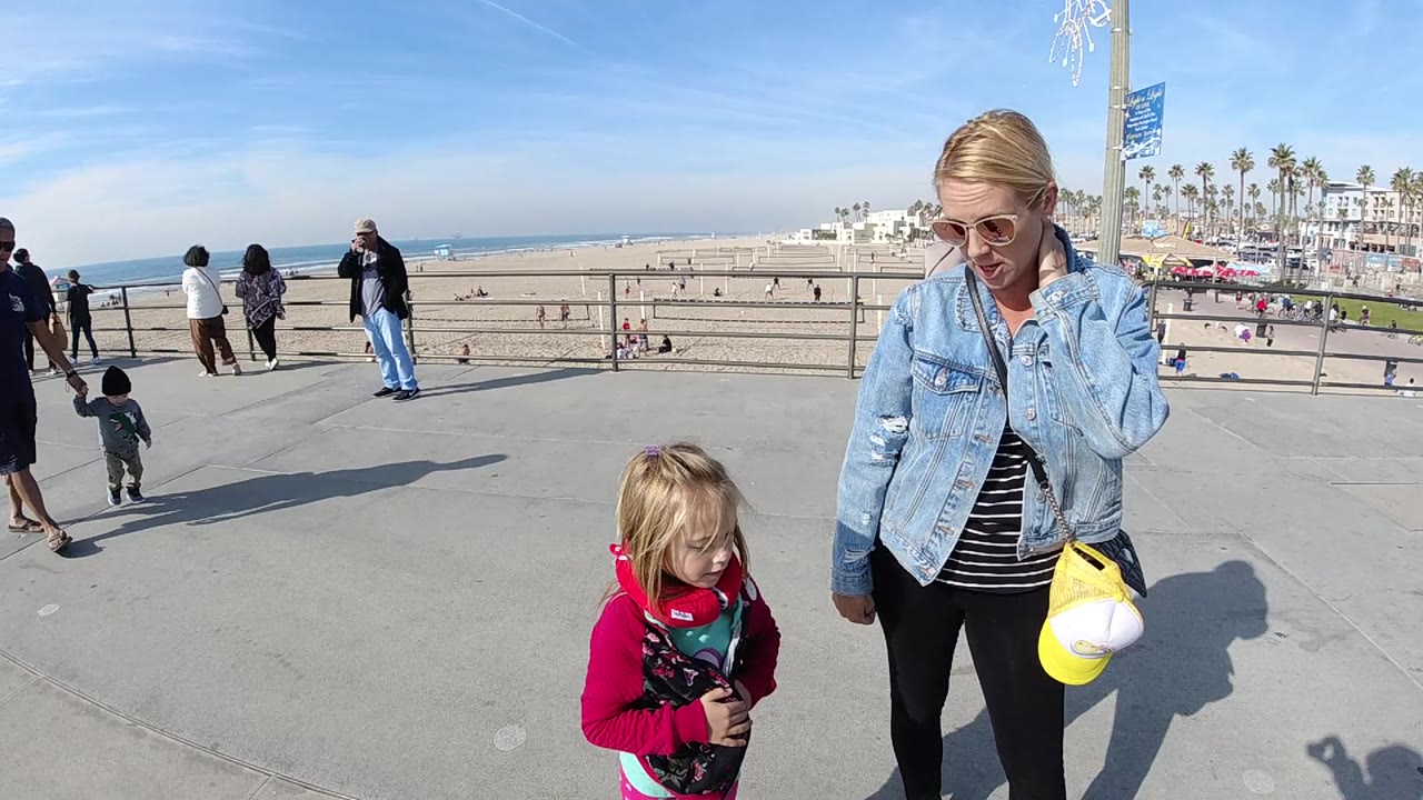 At 0:00 in Huntington Beach, a woman in a blue denim jacket stands beside a young girl in red on a concrete beach walkway with sand, ocean, pedestrians, and palm trees behind them.