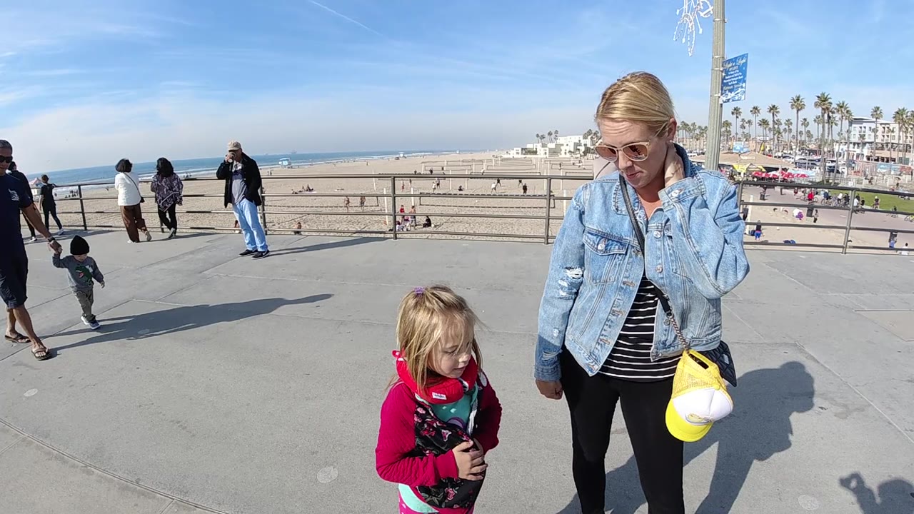 At 0:02 in Huntington Beach, the child stands slightly lower in frame while the woman remains at right, with palm-lined beachfront buildings and scattered people visible across the background.