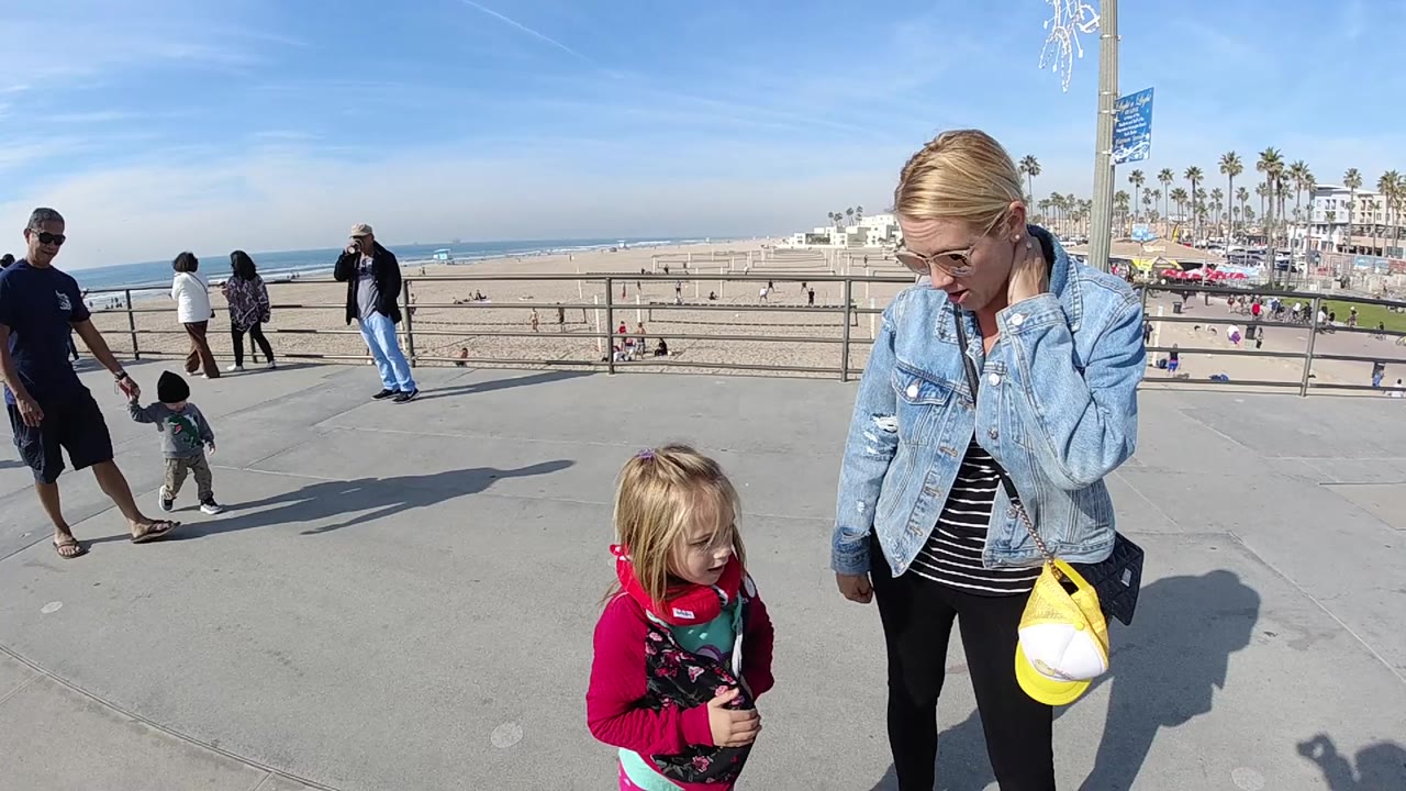 At 0:00 in Huntington Beach, a woman in a denim jacket stands beside a young girl in pink on a sunny concrete promenade, with the beach, ocean, railings, pedestrians, and palm trees behind them.