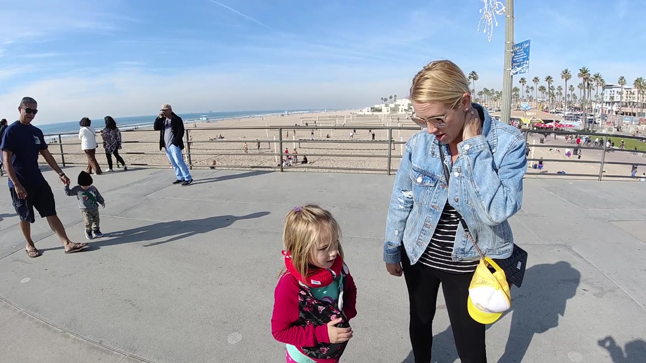 At 0:02, the child in a pink top stands lower in frame while the woman in denim holds a yellow item, with Huntington Beach sand, surf, palms, and distant buildings in the background.