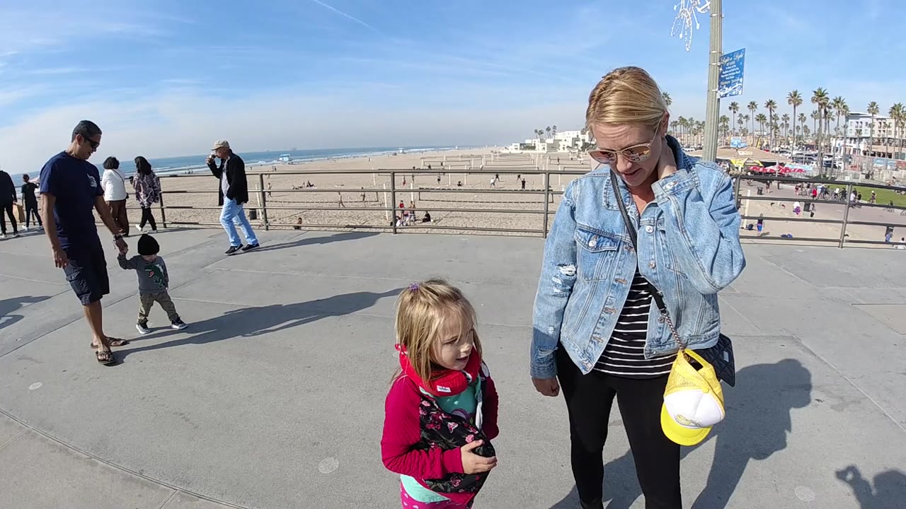 At 0:01 in Huntington Beach, the young girl turns slightly on the concrete promenade as the woman looks downward, with beachgoers, railings, sand, and ocean beyond them.