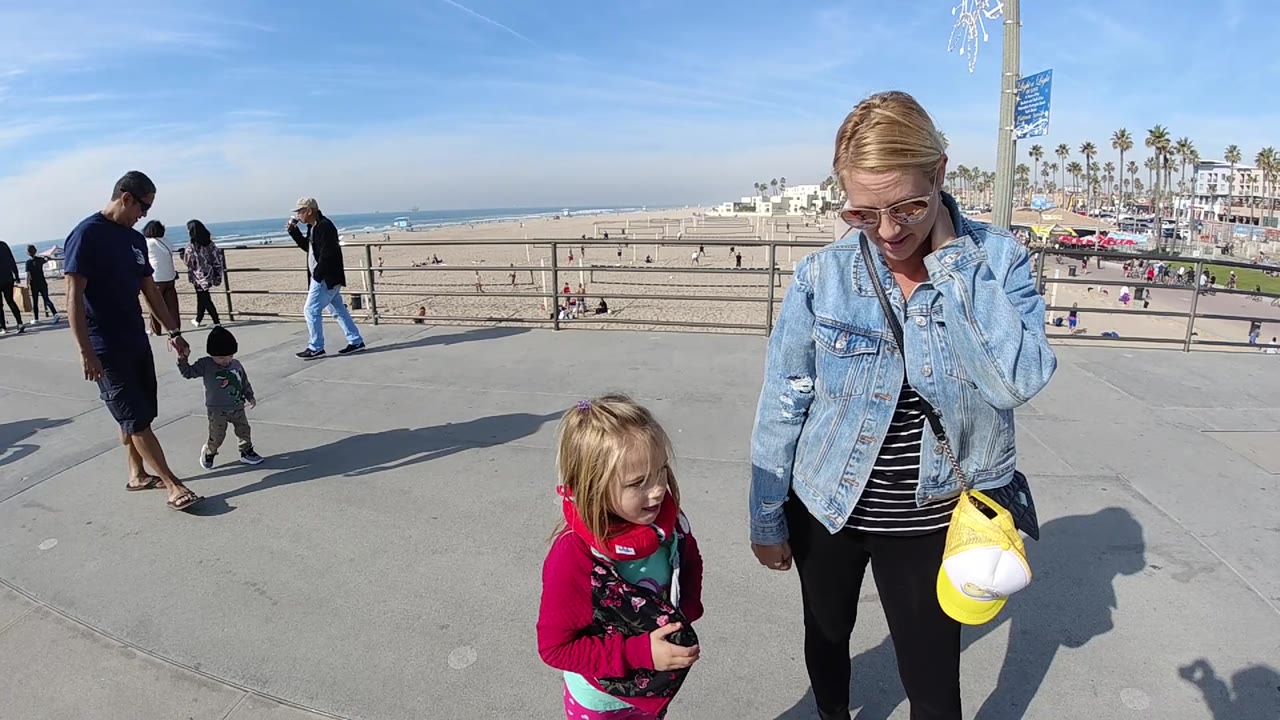 At 0:02 in Huntington Beach, the woman and child hold the foreground while a man and small child walk on the left and the ocean horizon stays visible past the railing.