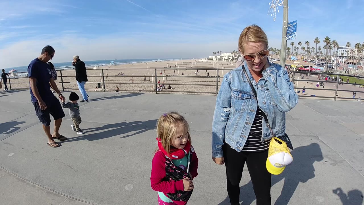 At 0:01, the woman and girl remain in the foreground on the Huntington Beach walkway while the man and small child stand near the rail to the left.
