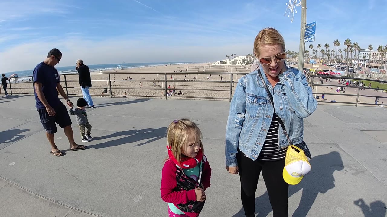 At 0:02, the girl in pink stands slightly ahead of the woman on the Huntington Beach promenade, with beachgoers spread across the sand below.
