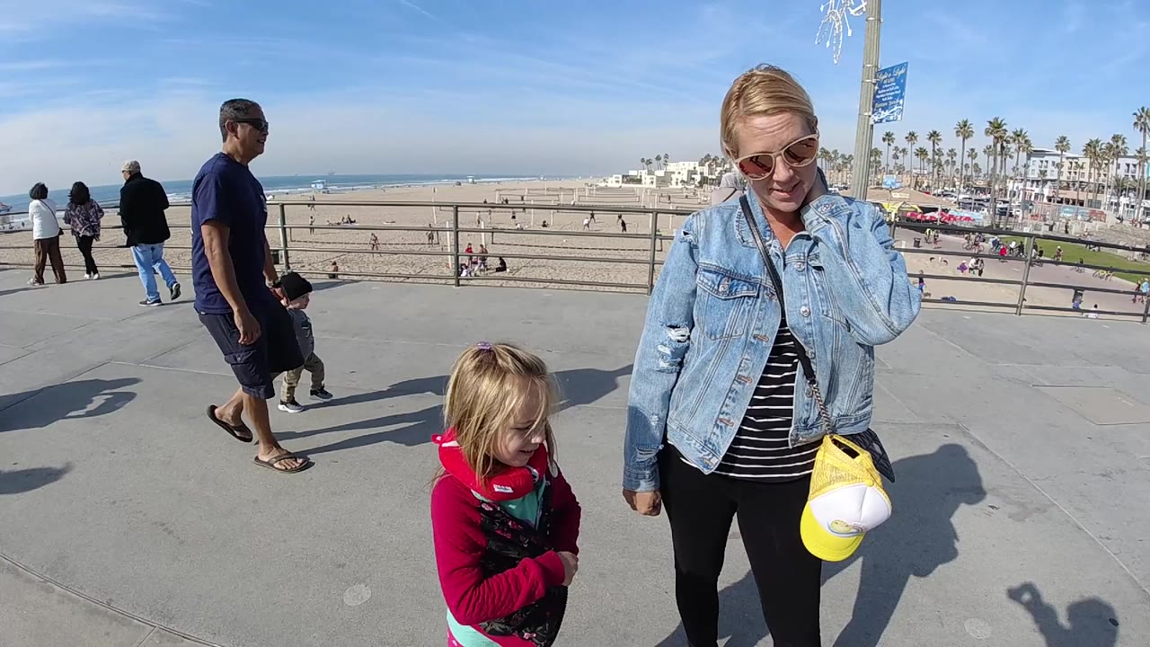 In Huntington Beach, California, a woman in sunglasses and denim stands beside a young girl on a concrete promenade with beach, ocean, palms, and pedestrians behind them.