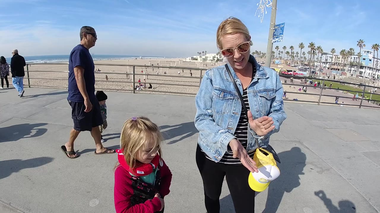 At 0:01, the woman looks down toward her hands and the yellow and white cap while the child stands in the foreground on the Huntington Beach walkway.