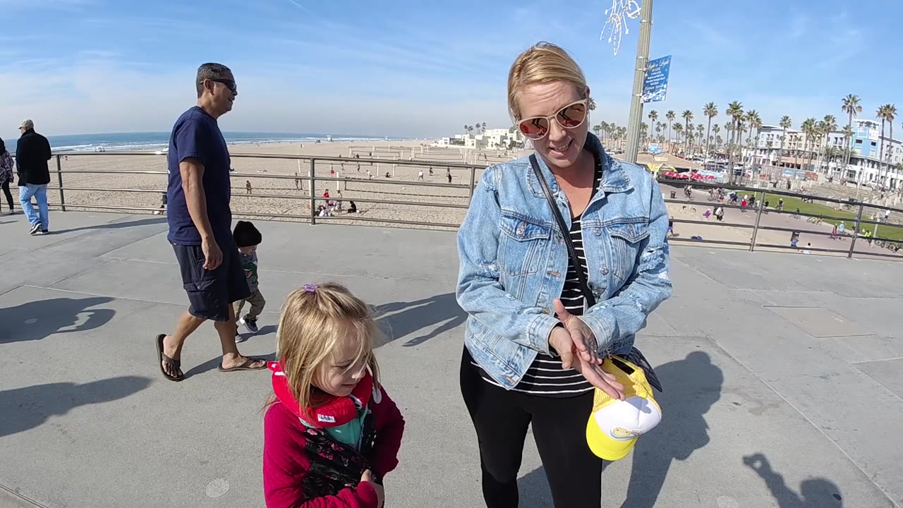 At 0:02, the woman and child stay in the foreground while a man walks behind them and the Huntington Beach sand, ocean, and palm-lined street remain visible.