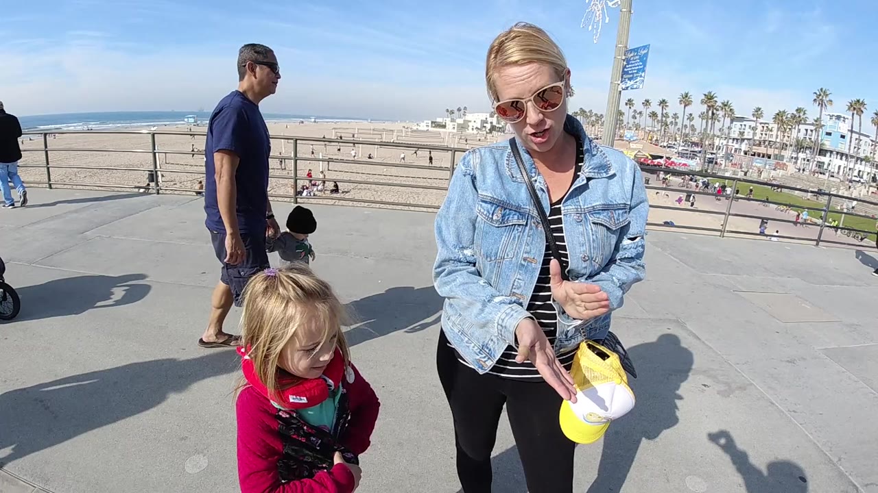 At 0:01 in Huntington Beach, the woman faces the camera while clapping, the little girl stands low in the foreground, and a toddler and walking man appear near the railing with the beach beyond.