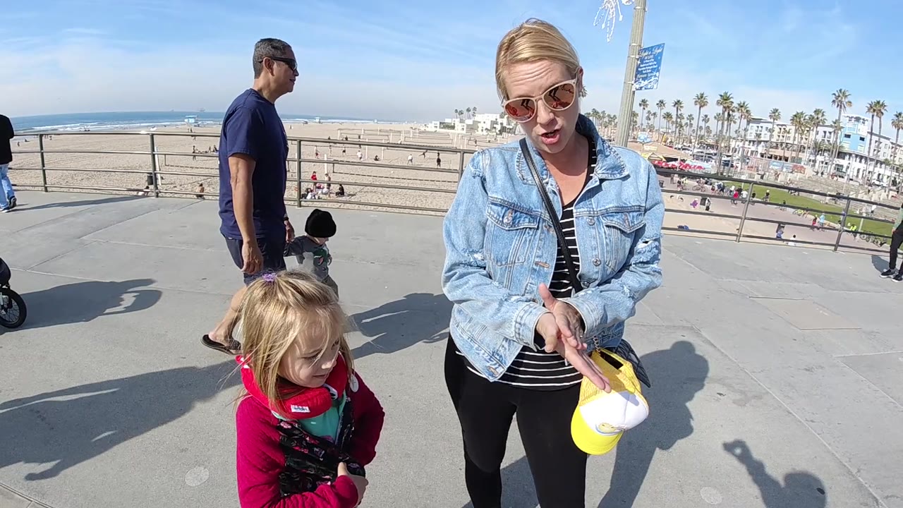 At 0:02 in Huntington Beach, the woman lowers her hands toward a yellow-and-white cap, a little girl stands beside her, and the toddler and man are visible near the beach railing.