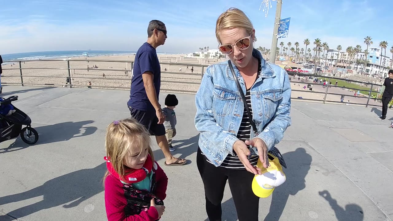 At Huntington Beach, California, a woman in sunglasses holds a yellow hat while standing beside a toddler in a red carrier near a beach railing, with sand, ocean, palms, and pedestrians behind them.
