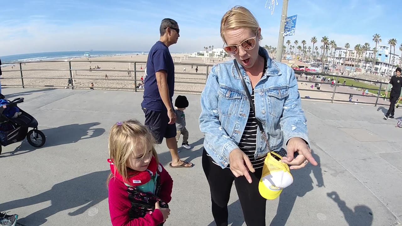 The woman begins extending her right arm and points downward or off to the side as the toddler stays near her, with bright concrete, railing shadows, beach sand, ocean, and palm-lined buildings behind them.