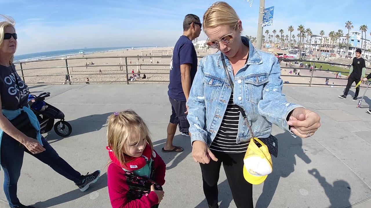 At Huntington Beach, a woman in a denim jacket stands near a small child in red while other pedestrians pass along a paved overlook above the sand and ocean.