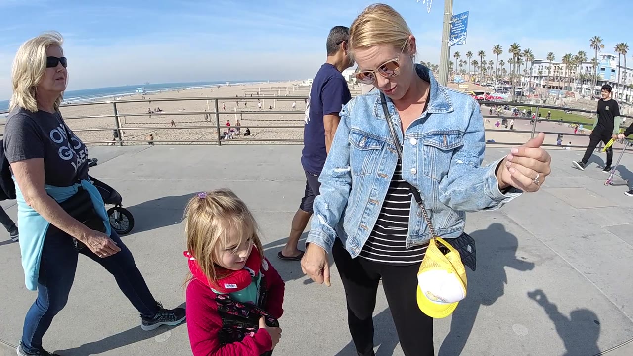 At Huntington Beach, the child stands lower in frame while the woman holds a yellow cap, and several pedestrians move along the oceanfront walkway behind them.